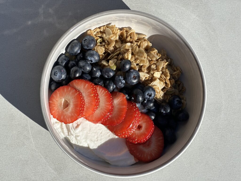 The image shows a bowl of Greek yogurt topped with Bodacious granola and fresh berries as part of the Blue Zone day of eating.