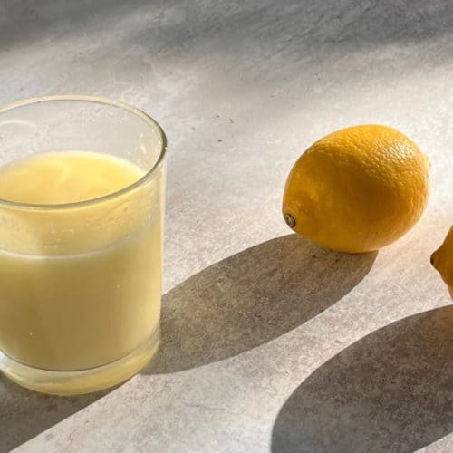 Freshly squeezed lemon juice in a glass with two whole lemons on a light-colored surface under natural sunlight.