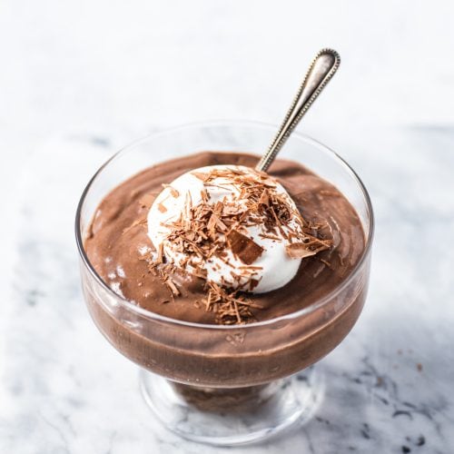 Rich chocolate mousse topped with whipped cream and chocolate shavings in a glass dessert cup on a white marble surface. Perfect for chocolate lovers and dessert enthusiasts.