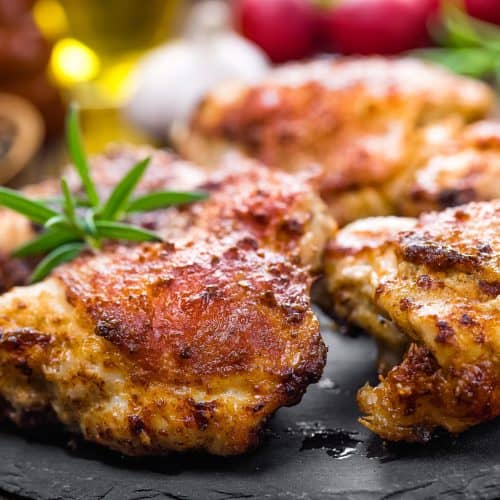 Juicy grilled chicken thighs with crispy, golden-brown skin garnished with fresh herbs on a black slate serving platter. Perfect for barbecue dinners and savory meal options.