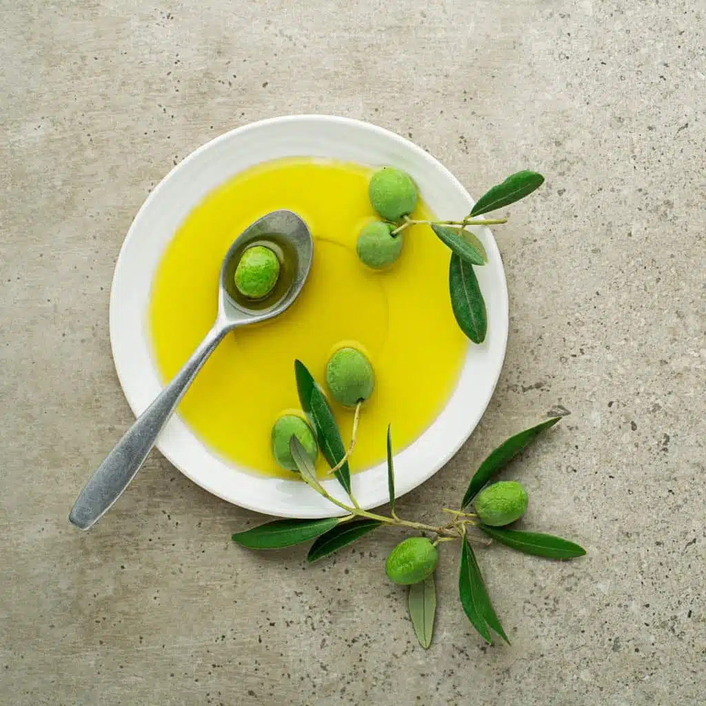 Fresh green olives in olive oil with a silver spoon and olive branch on a neutral background.