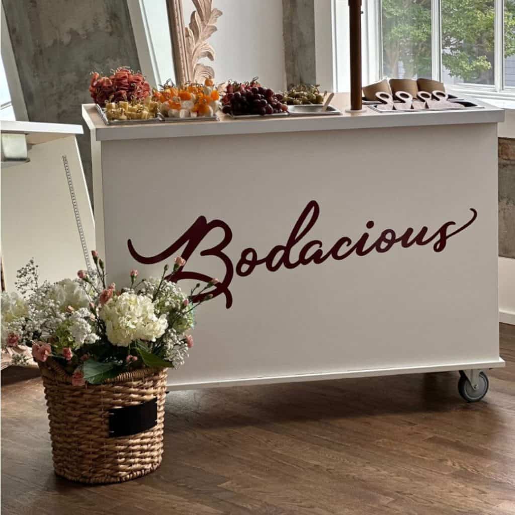 Bright white Bodacious Shops mobile beverage cart with elegant burgundy logo, displayed with fresh fruit and floral arrangements, ideal for events and catering.